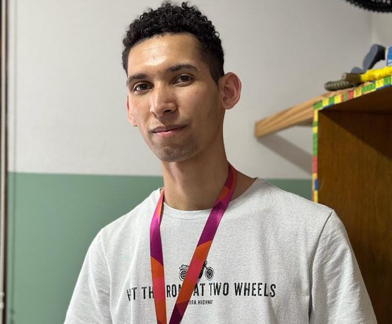 Na imagem, temos Daniel, jovem branco com cabelos encaracolados bem curtos, ele veste uma camiseta branca e um crachá de identificação vermelho.